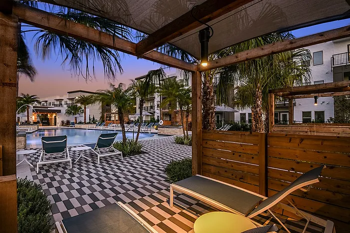 Luxurious apartment community with a serene poolside view from a cabana, featuring modern lounge chairs, palm trees, and a chic patterned tile patio at sunset.