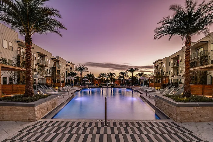 Luxurious apartment community featuring a stunning swimming pool lined with palm trees and modern lounge chairs at sunset.