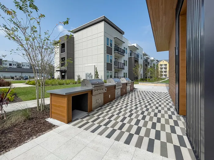 Modern apartment community with a stylish outdoor grill area and checkerboard flooring, surrounded by lush greenery.