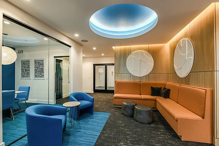 Modern clubhouse lounge area with vibrant orange seating, elegant blue accent chairs, and stylish wall art, illuminated by unique ceiling lighting.