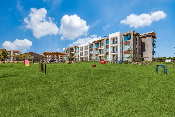 Contemporary apartment complex with spacious green dog park, agility equipment, and outdoor seating, set against a bright blue sky.