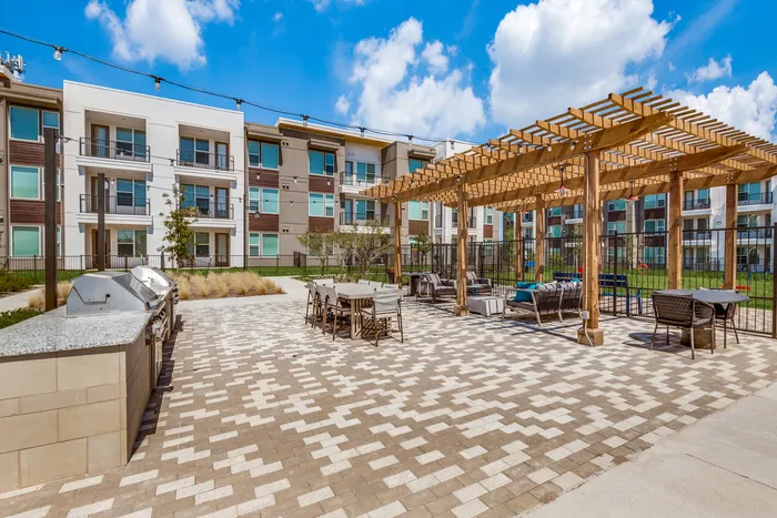 Vibrant courtyard view of modern apartment complex with outdoor grilling area, stylish seating under a wooden pergola, and lush green landscaping.