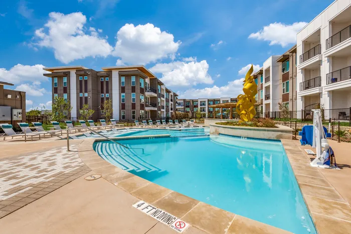 Luxurious apartment complex featuring a sparkling swimming pool, modern sun loungers, and artistic sculpture under a sunny blue sky.
