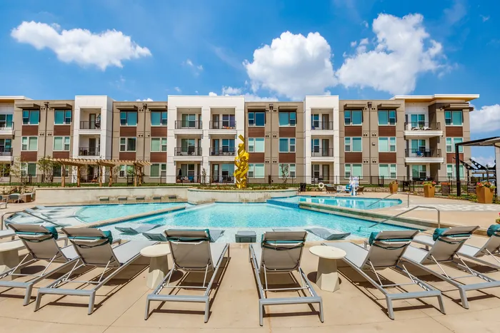 Modern apartment complex with a sparkling pool and sun loungers under a clear blue sky, perfect for relaxation and luxury living.