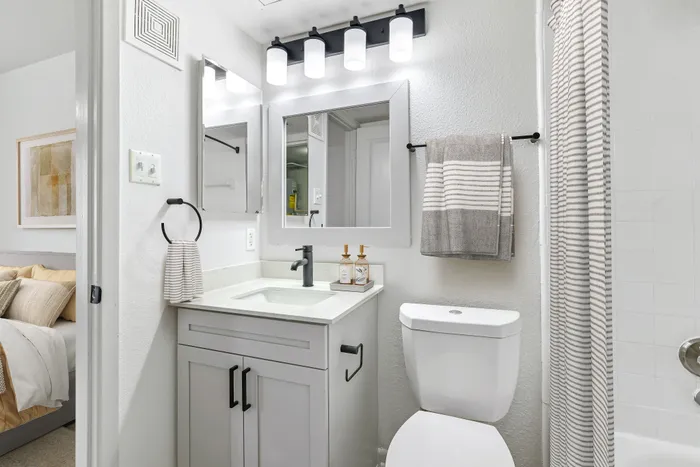 Bathroom with vanity mirror and lighting, looking into bedroom with tan bedding and wall art.