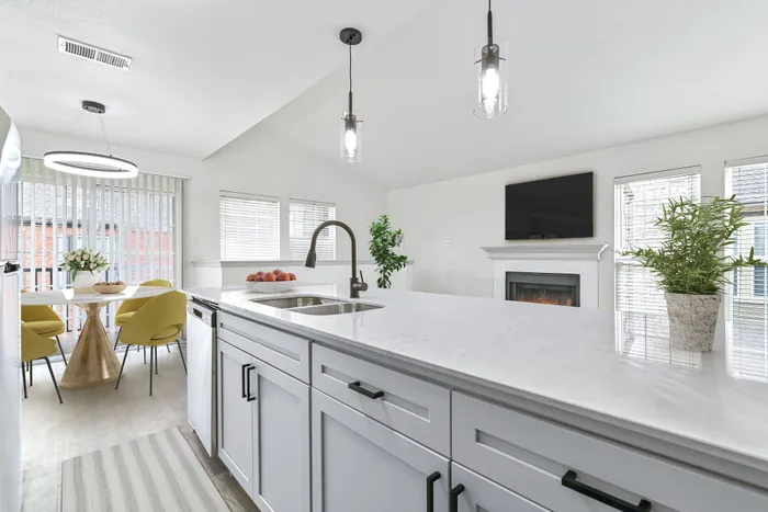 Kitchen with quartz counters, undermount sink and gooseneck faucet, pendant lighting, with living room in background with fireplace, large windows, and TV.