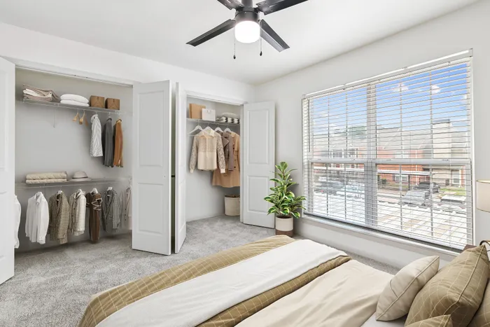 Carpetted bedroom with large center window, dual closets with clothing, queen bed with tan bedding, a ceiling fan and potted plant.