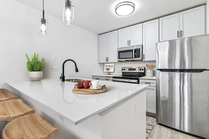 Kitchen with stainless appliances, kitchen bar with sink, pendant lighting, wooden barstools, and white cabinets.