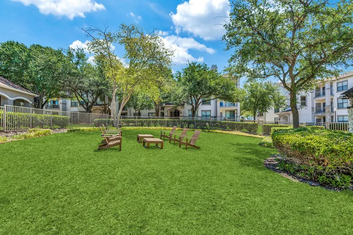 Beautifully landscaped outdoor space with cozy wooden seating, lush green lawn, and elegant apartment buildings surrounded by mature trees.
