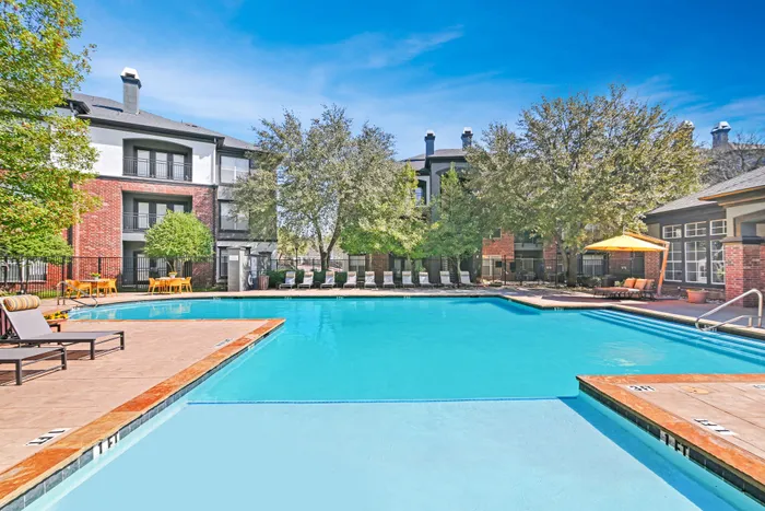 Large community pool with sundeck and chairs, brick building with trees