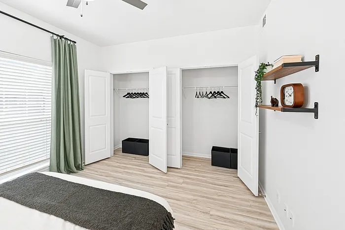 Bedroom with wooden flooring, double closets, large center window with green curtain, two floating shelves, queen bed with white and black bedding..