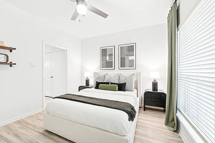 Bedroom with wooden floor, ceiling fan, wall art, large window, black side table with lamp, and floating shelves.