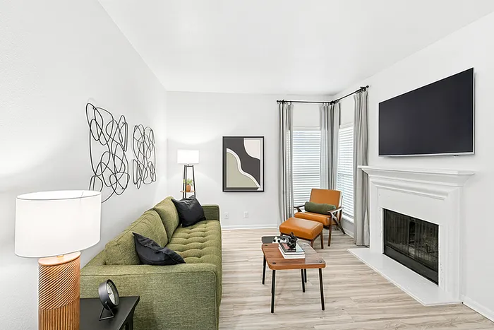 Living room with sofa, side table with lamp, wooden coffee table, fire place, wall mounted TV, two windows and wall art.