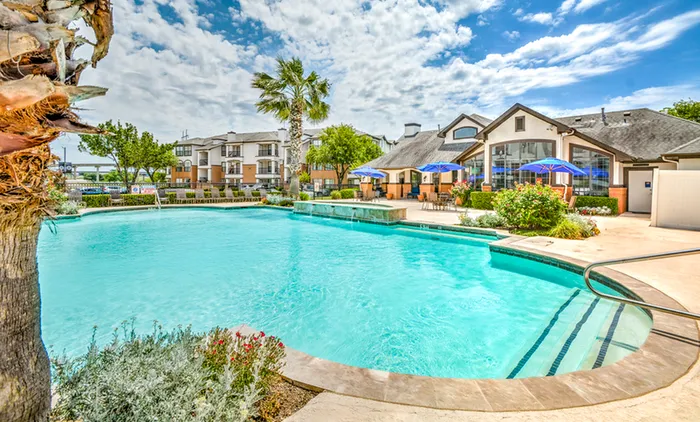 Luxurious apartment complex featuring a sparkling pool, lush landscaping, and vibrant patio umbrellas under a clear blue sky.