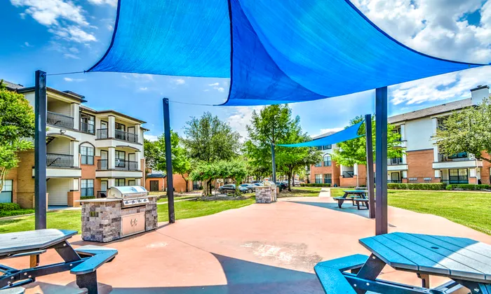 Charming outdoor communal area featuring modern shade sails, picnic tables, and a state-of-the-art BBQ grill surrounded by lush greenery and elegant apartment buildings.