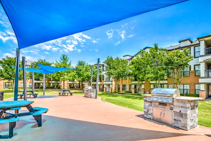Vibrant outdoor patio area with blue shade sails, picnic tables, and a modern barbecue grill surrounded by lush greenery and stylish apartment buildings.