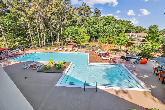 Apartment community featuring a swimming pool, surrounded by sun loungers, bright umbrellas, and lush greenery for a relaxing retreat.