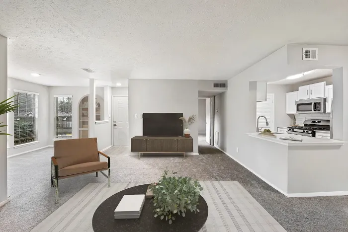 “Open-concept apartment living room with plush carpeting, modern furnishings, large windows, and breakfast bar overlooking a kitchen with stainless steel appliances.