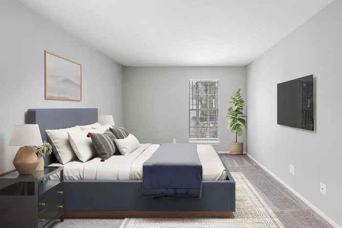 Spacious apartment bedroom with plush carpeting, modern furnishings, large window, and wall-mounted TV in a neutral color palette.