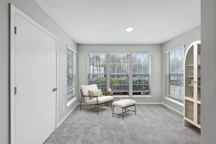 Bright apartment sunroom or flex space with plush carpeting, large windows for natural light, and cozy seating area.