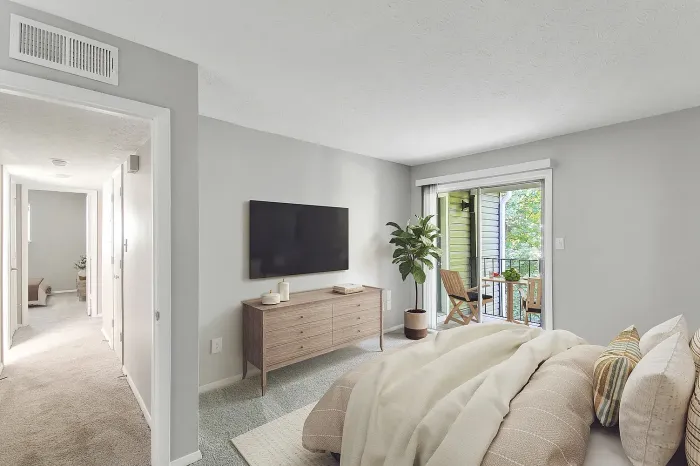 Spacious apartment bedroom with plush carpeting, wall-mounted TV, sliding glass door to private balcony, and natural light.