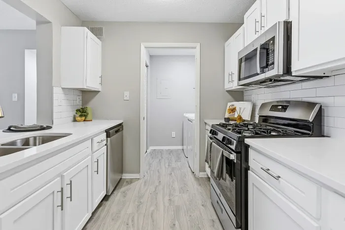 Modern galley-style apartment kitchen with stainless steel appliances, quartz countertops, white shaker cabinets, subway tile backsplash, and adjacent laundry area.