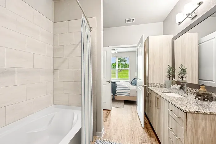 Modern bathroom with elegant tiled shower and granite countertop, offering a glimpse into a cozy, well-lit bedroom with lush green views beyond.