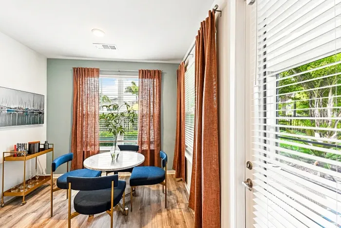 Bright, modern dining nook with a round white table, four blue upholstered chairs, and warm rust-colored curtains framing large windows and a glass door, letting in natural light and views of greenery outside.