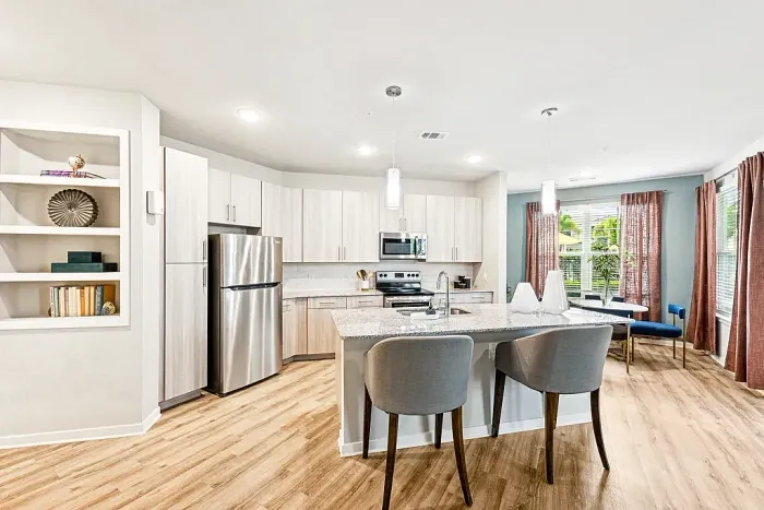 Bright, modern kitchen with stainless steel appliances, light wood cabinetry, and a granite island with bar seating, adjacent to a cozy dining area with blue chairs, a round table, and large windows with rust-colored curtains.