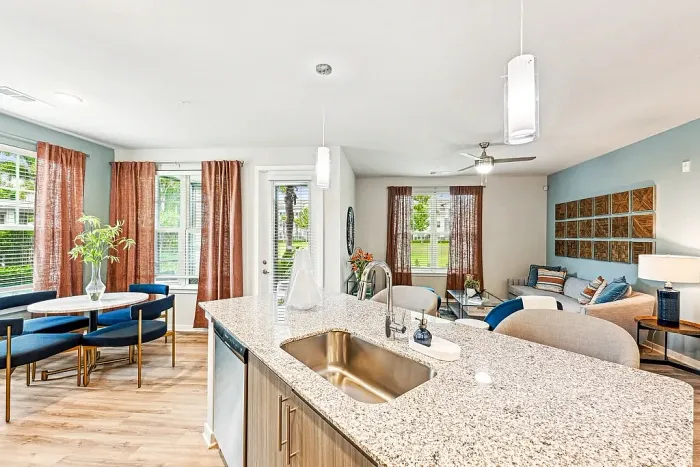 Open-concept apartment featuring a modern kitchen with a granite island and sink, flowing into a bright dining area with blue chairs and a cozy living room with a sofa, wall art, and large windows with rust-colored curtains.