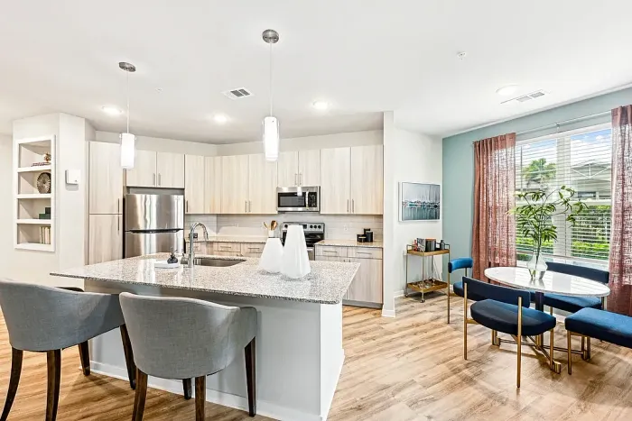 Bright, modern kitchen with stainless steel appliances, light wood cabinetry, and a granite island with bar seating, adjacent to a cozy dining area with blue chairs, a round table, and large windows with rust-colored curtains.