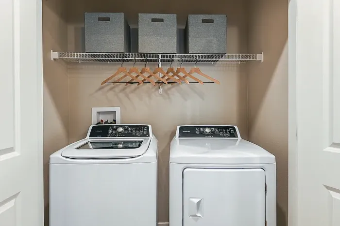 Compact laundry closet with a top-load washer and front-load dryer, overhead wire shelving with storage bins, and a hanging rod with wooden hangers.