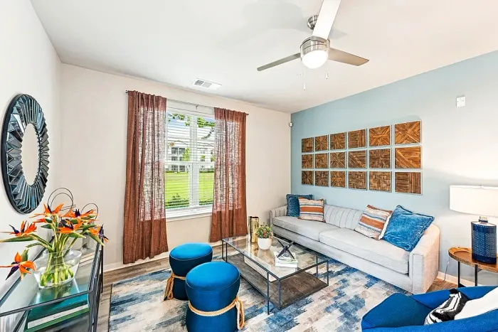 Bright, stylish living room with a gray sofa, blue accent pillows and ottomans, a glass coffee table, and decorative wall art, with natural light from a window framed by rust-colored curtains.