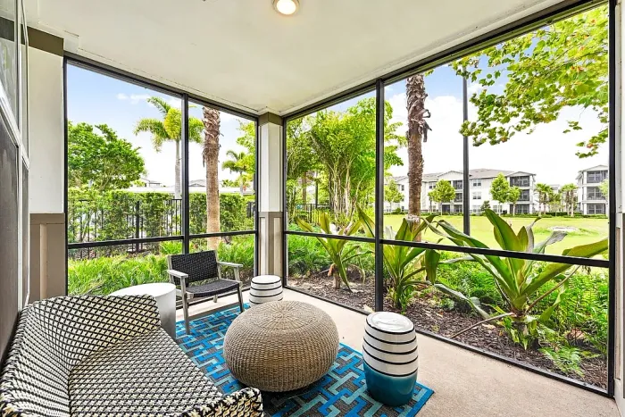 Screened-in patio with comfortable seating, including a lounge chair, accent stools, and a woven ottoman, overlooking lush landscaping with palm trees.