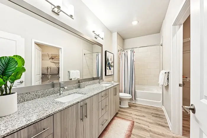 Spacious bathroom with a double-sink vanity, granite countertops, large mirror, and wood-style cabinetry, leading to a tub-shower combo with a curtain and adjacent walk-in closet.