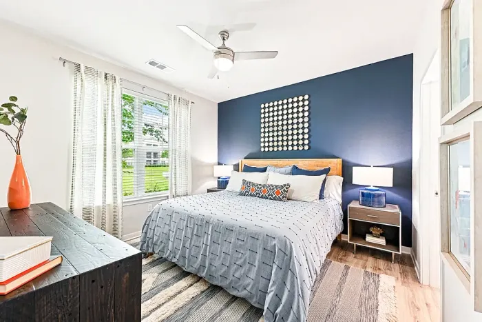 Bright bedroom with a queen bed, navy accent wall, modern decor, and matching nightstands with lamps, complemented by a large window with sheer curtains and natural light.