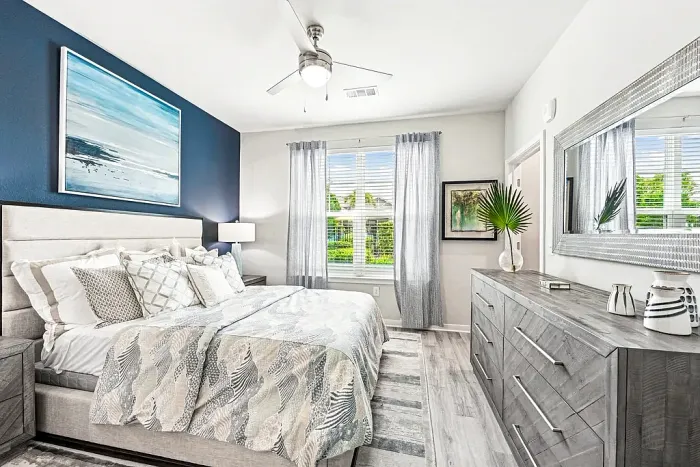 Bright, modern bedroom with a large bed, blue accent wall, and coordinated decor, featuring a dresser with a mirror, ceiling fan, and a window with sheer curtains allowing natural light.