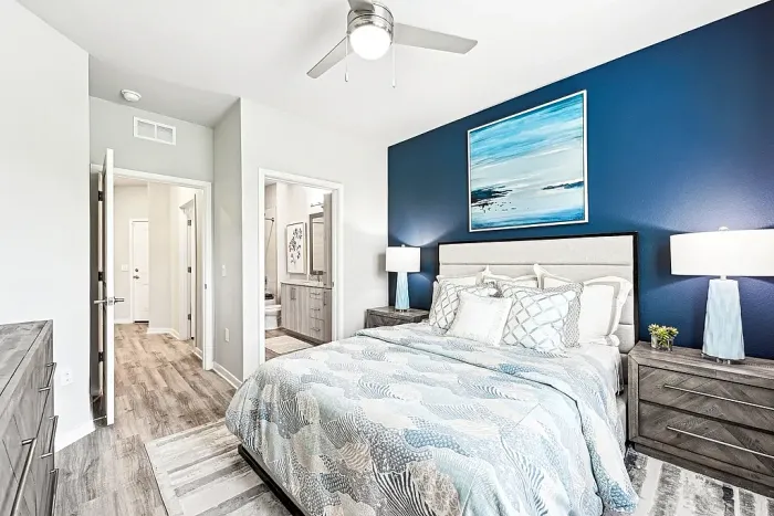 Spacious bedroom with a queen bed, blue accent wall, and coordinated decor, featuring matching nightstands and lamps, with an open view to an en-suite bathroom and hallway.