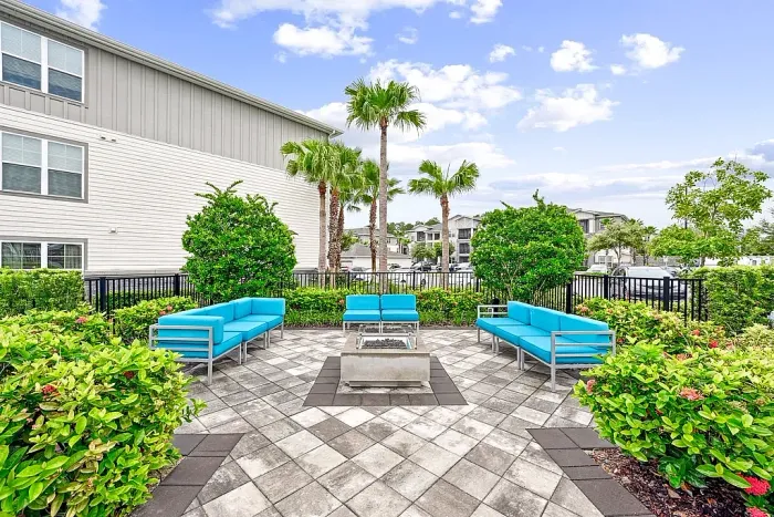 Outdoor community patio with modern seating, including teal cushioned sofas and chairs arranged around a central fire pit, surrounded by lush landscaping and palm trees.