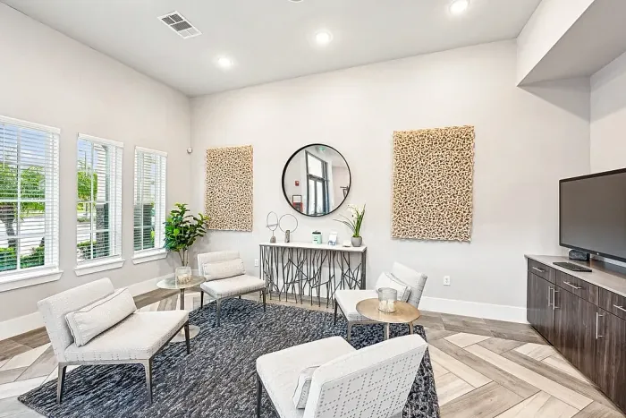 Bright clubhouse lounge area with modern seating, accent chairs, and small tables arranged on a textured rug, featuring decorative wall art, a console table with a round mirror, and a TV.