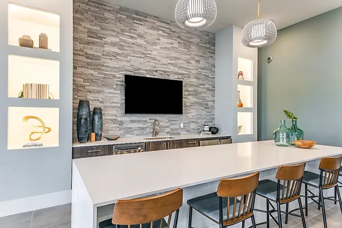Modern clubhouse kitchen and bar area with a large island, bar seating, pendant lighting, and a stone accent wall with a mounted TV and built-in shelving.