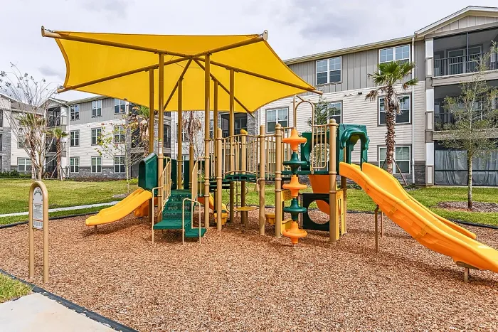 Community playground with slides and climbing structures under a bright shade canopy, set within a landscaped apartment complex.