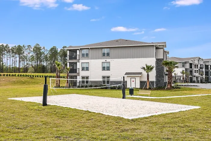 Sand volleyball court with net set in a grassy open area, surrounded by apartment buildings and landscaped grounds.