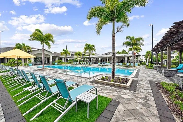 Resort-style swimming pool with lounge chairs, umbrellas, and palm trees, set within a landscaped apartment community with spacious deck seating.