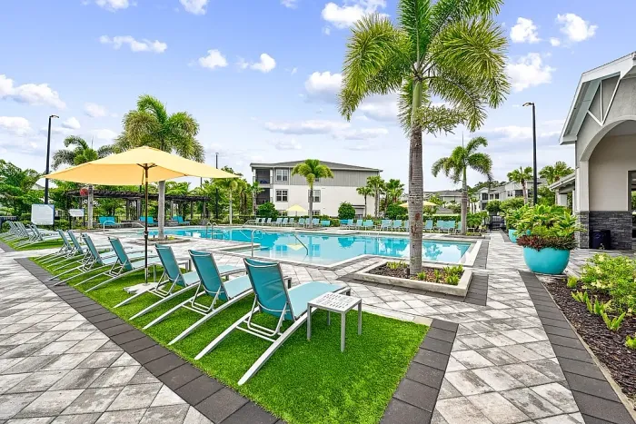 Resort-style swimming pool with lounge chairs, umbrellas, and palm trees, set within a landscaped apartment community with spacious deck seating.