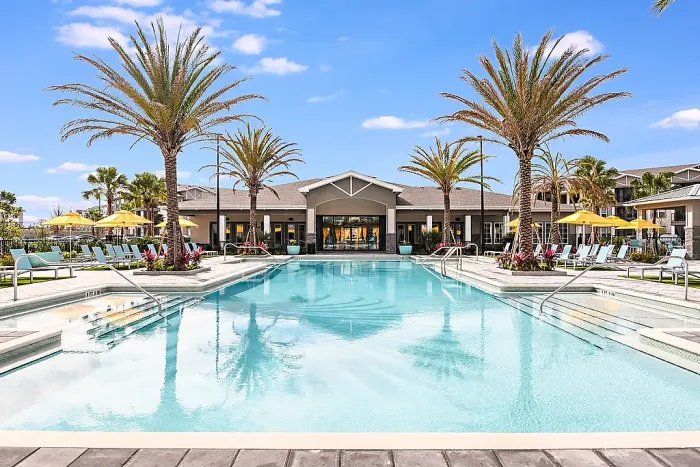 Large resort-style pool with shallow entry areas, surrounded by lounge chairs, umbrellas, and palm trees, with a clubhouse in the background.