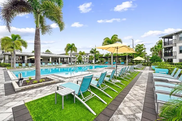 Resort-style swimming pool with lounge chairs, umbrellas, and palm trees, set within a landscaped apartment community with spacious deck seating.