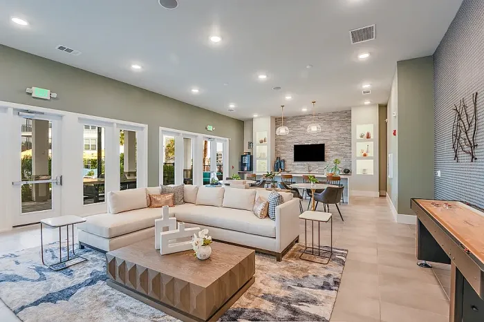 Spacious clubhouse lounge with a sectional sofa, coffee table, and open-concept layout leading to a dining area and kitchen, featuring modern decor, built-in shelving, and a game table.