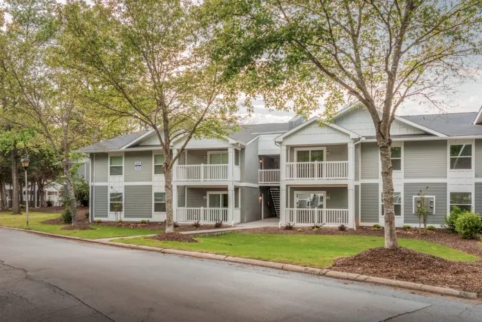 Inviting grey and white apartment complex with lush green landscaping, featuring charming balconies and surrounded by mature trees.