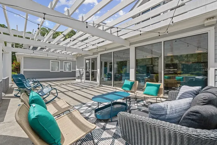 Outdoor patio lounge with cushioned seating, lounge chairs, and a pergola overhead with string lights, adjacent to large glass doors leading into the clubhouse.